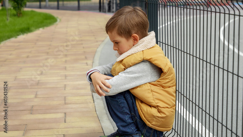 Vászonkép Unhappy lonely boy leaning on metal fence at school playground and sitting down