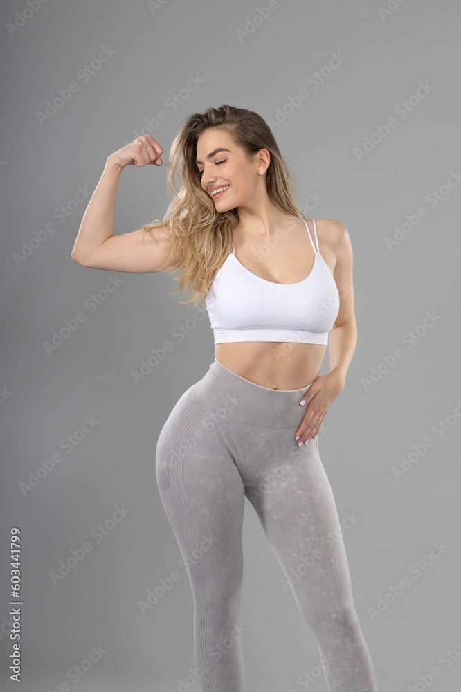 Fototapeta premium Young white fitness woman in sportswear posing in studio with gray background, showing biceps and smiling.beautiful attractive young girl with natural makeup and loose hair posing in studio