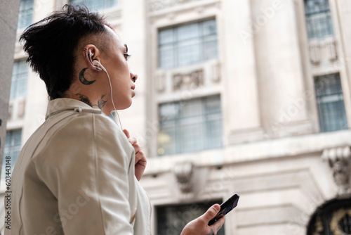 Androgynous male is using headphones to listening to music in front of building.