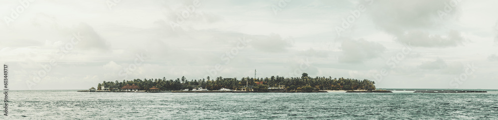Maldives; Panorama of a Maldivian islet featuring lush green trees, surrounded by the vast calm blue sea, grey clouds in the sky contrast with the overall tranquil scene