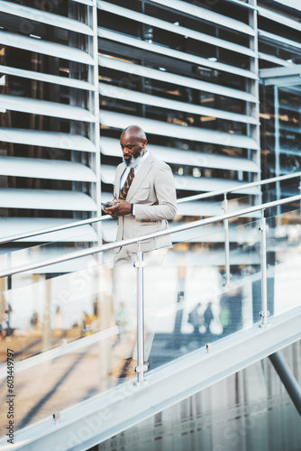 A sleekly dressed bald businessman in a beige suit and striped tie is texting on his smartphone while walking down the glass seamless railing stairs in Lisbon