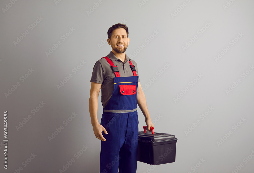 Portrait of a happy smiling confident repairman with beard holding a tool box in hands isolated on gray wall background. Professional young handyman worker standing cheerful in blue overall. Portrait of a happy smiling confident repairman with beard holding a tool box in hands isolated on gray wall background. Professional young handyman worker standing cheerful in blue overall.