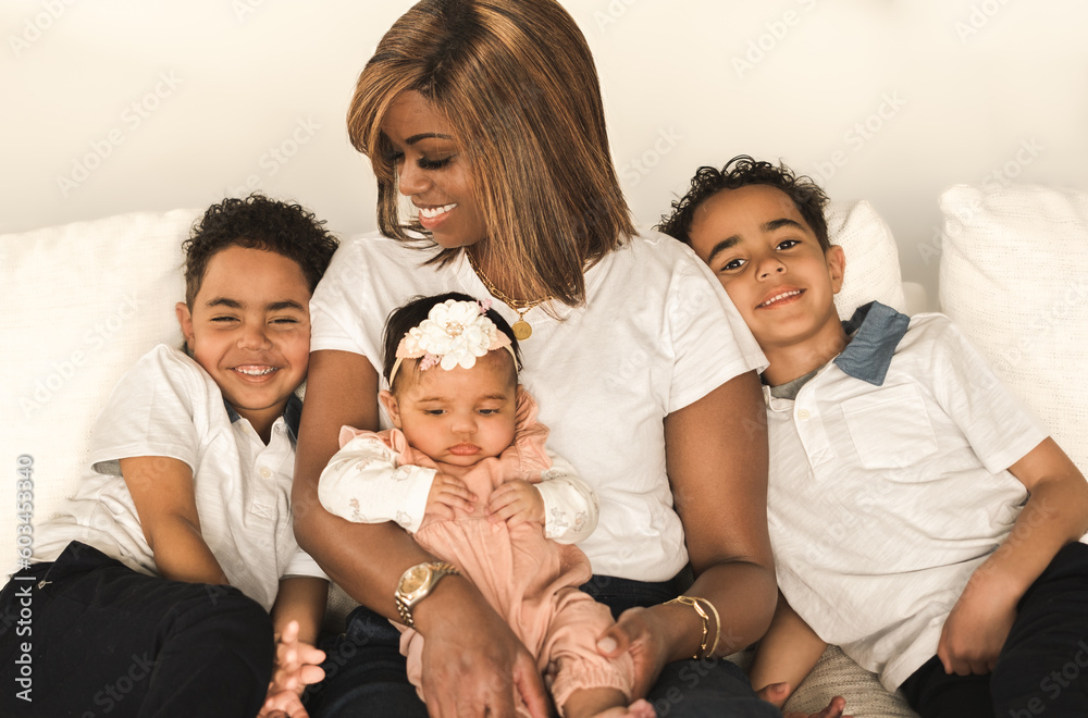 View of beautiful African American woman sitting on couch with her smiling six year old twin boys and baby daughter on her lap; light wall in background View of beautiful African American woman sitting on couch with her smiling six year old twin boys and baby daughter on her lap; light wall in background