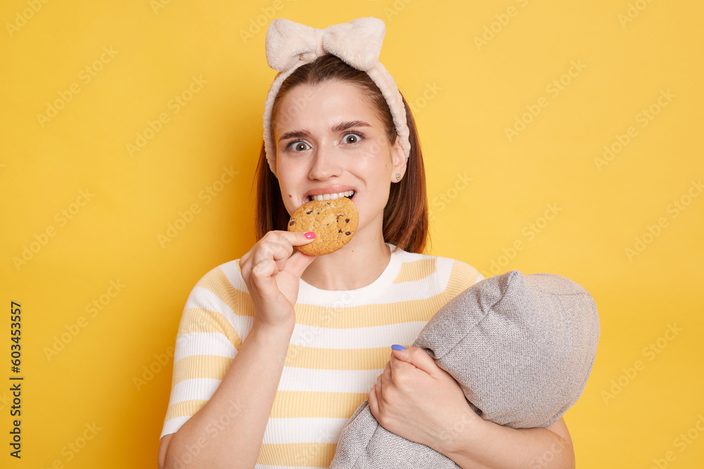 Confused woman wearing striped T-shirt and hair band standing isolated over yellow background posing with pillow in hands biting delicious cookie looking at camera. Confused woman wearing striped T-shirt and hair band standing isolated over yellow background posing with pillow in hands biting delicious cookie looking at camera.