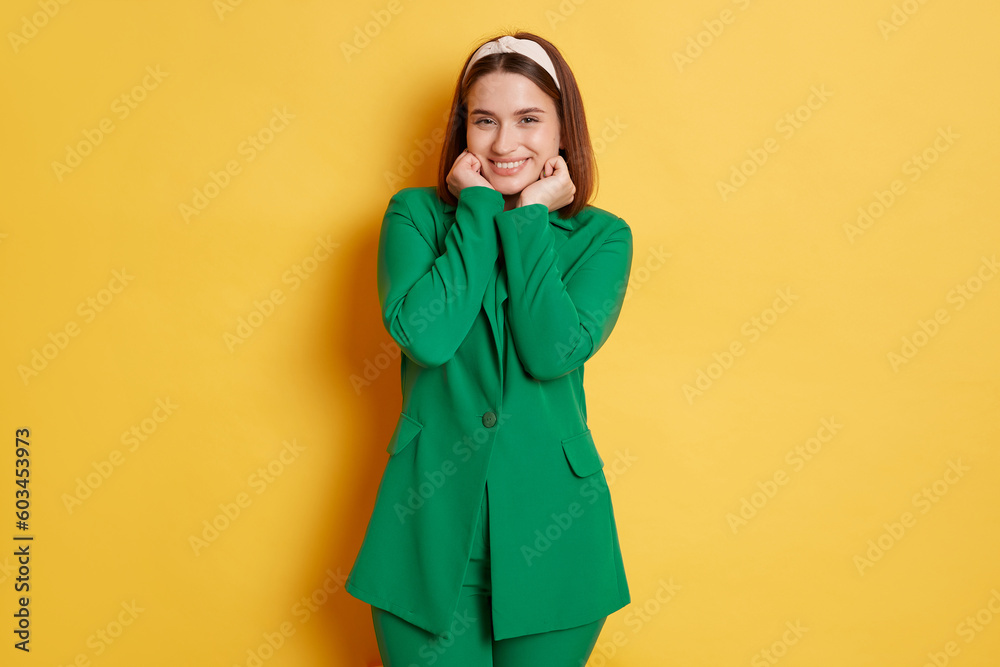 Smiling happy attractive woman wearing green jacket keeps hands under chin looking at camera with cute sly expression standing isolated over yellow background. Smiling happy attractive woman wearing green jacket keeps hands under chin looking at camera with cute sly expression standing isolated over yellow background.
