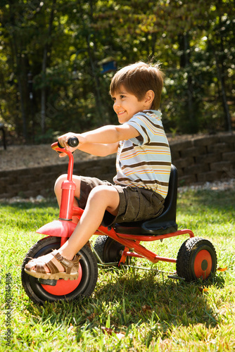 Side view of Hispanic boy riding red tricycle in grass.