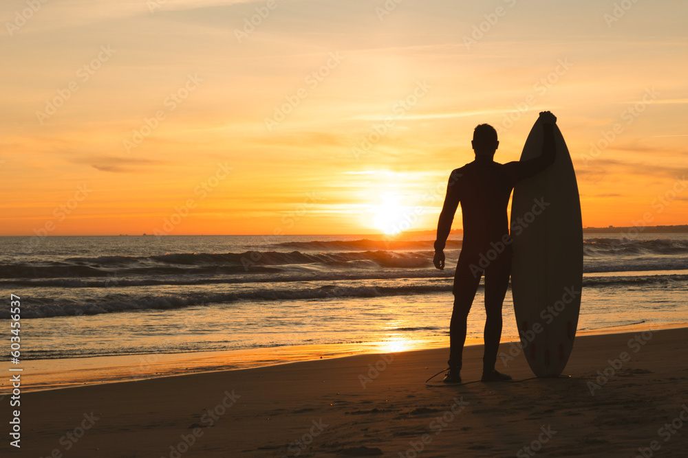 A fit man stands on the seashore leaning on a surfboard at sunset