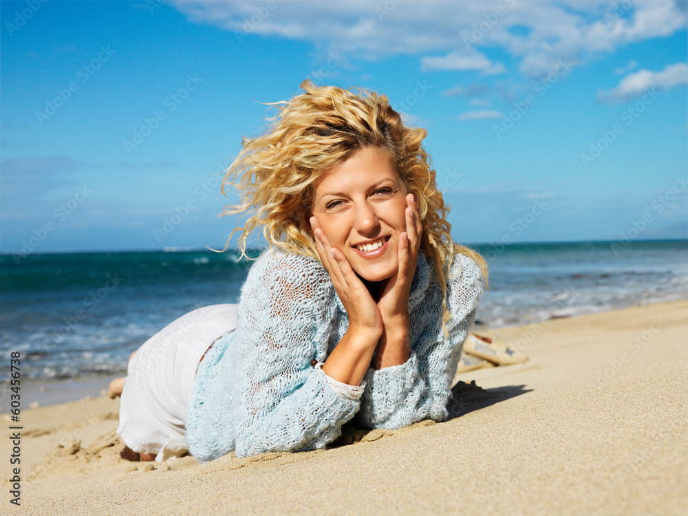 Pretty young blond woman lying in sand on Maui, Hawaii beach with head resting on hands smiling.
