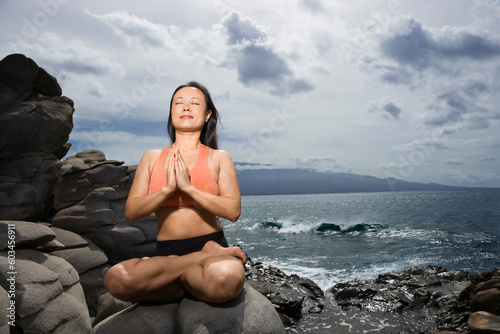 Asian woman sitting on rock by ocean in lotus pose with eyes closed in Maui, Hawaii © Designpics