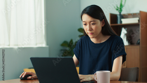 Beautiful young asian woman looking at screen, using laptop browsing the internet. Happy focused attractive lady typing laptop computer sits at desk in living room working online remotely from home © STAGE65