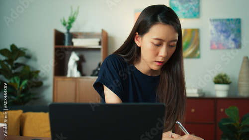 Beautiful young asian female freelancer looking at screen, typing, focusing on laptop. Attractive Asian woman sits at desk at living room working seriously while checking paperwork and taking notes.