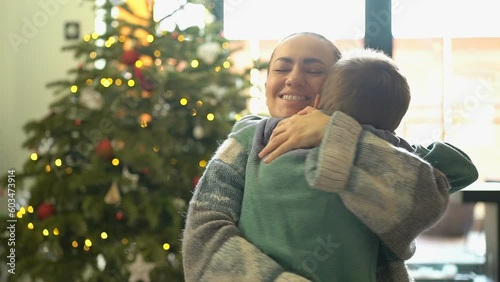 Portrait of mother and boy hugging in front of christmas tree. A little boy runs right into her arms.
