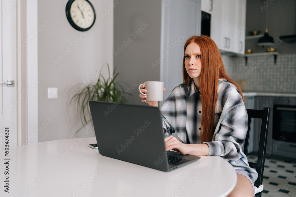 Fototapeta premium Portrait of thoughtful freelancer female remote working or studying on laptop computer sitting at table, holding in hand coffee cup, thinking looking away. Cute redhead young woman using laptop.