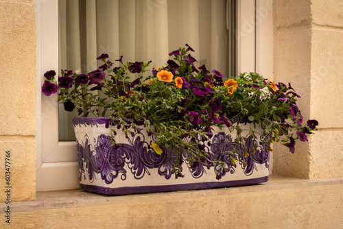 Purple and orange garden pansy flowers in a rectangular ceramic pot on a windowsill outside the building. 