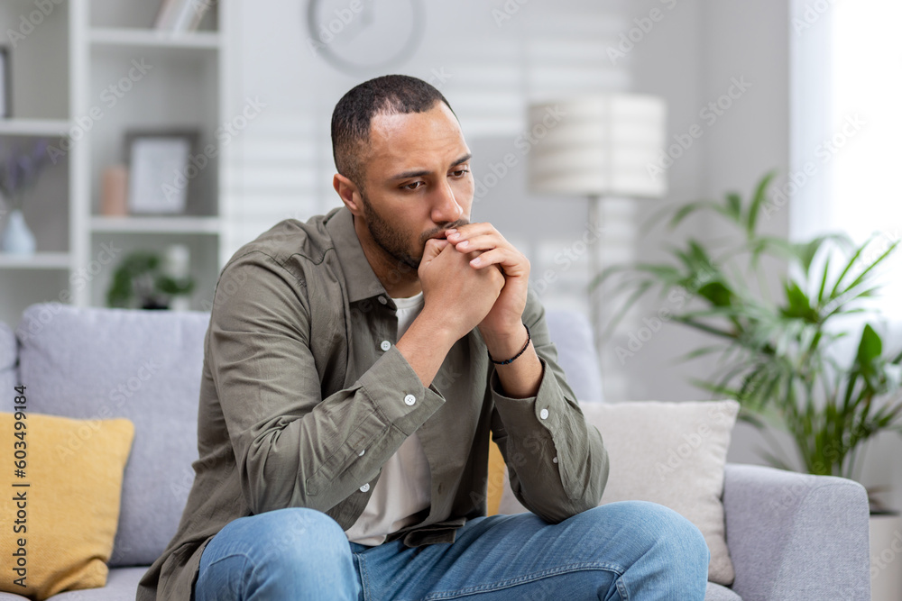 Upset man sitting alone at home, depressed hispanic man on sofa in living room in casual clothes thinking about difficult decision