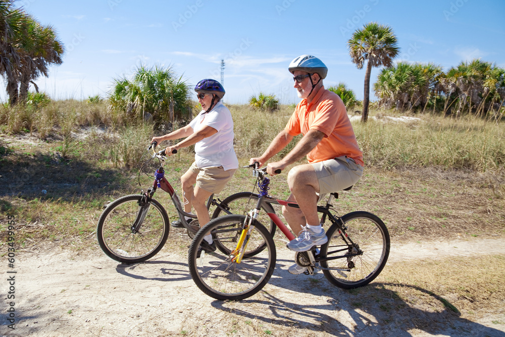 Senior couple riding bikes at the beach, wearing sunglasses and helmets. Focus on the woman. Senior couple riding bikes at the beach, wearing sunglasses and helmets. Focus on the woman.