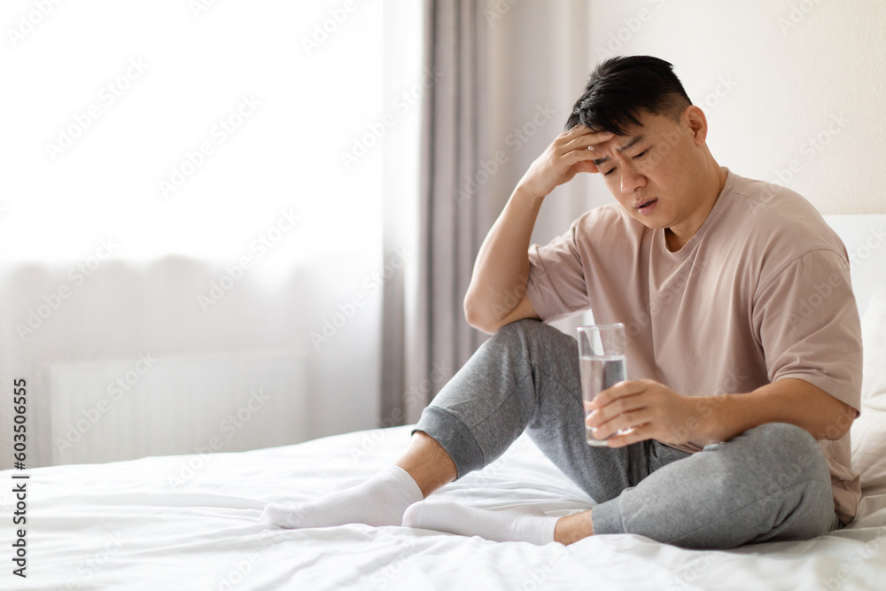 Asian man in pain sitting on bed, holding glass water Asian man in pain sitting on bed, holding glass water