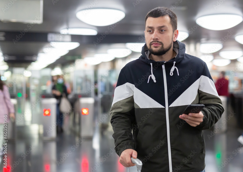 Obraz premium Interested young man browsing messages on phone while waiting for subway train, standing at station..