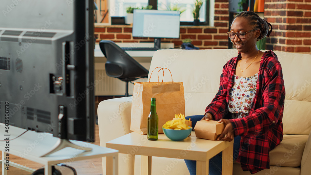 Young adult eating cheeseburgers with fries and beer, having fun with comedy movie on television. Relaxed girl enjoying cheeseburger and snacks, binge watching show in living room. Young adult eating cheeseburgers with fries and beer, having fun with comedy movie on television. Relaxed girl enjoying cheeseburger and snacks, binge watching show in living room.