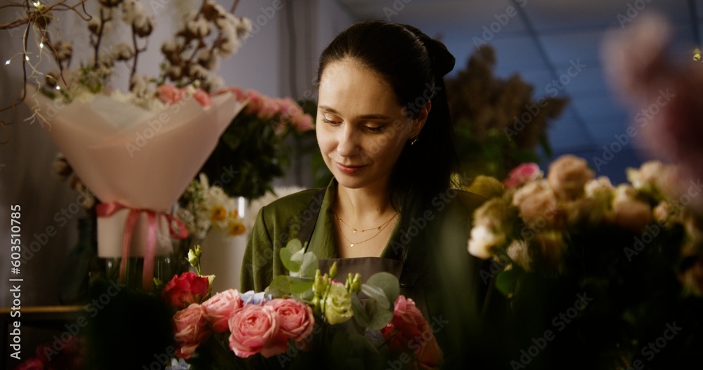 Professional female florist keeps in hands beautiful bouquet, smiles and looks at camera. Vases with plants and fresh flowers stand at background. Retail floral business and entrepreneurship concept.