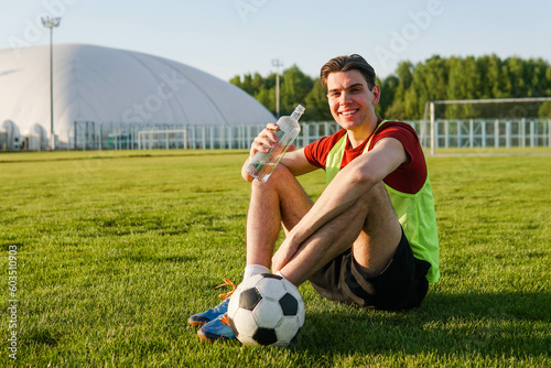 Young man football player resting after training sitting on grass and drinking water from a bottle. Soccer ball on the field