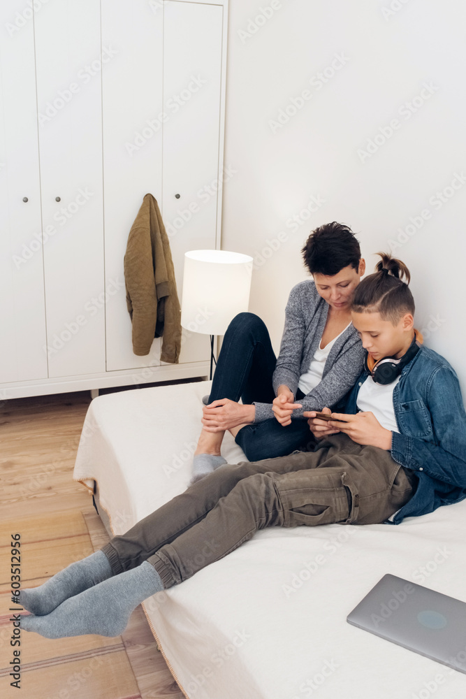 Teenager boy addicted to technology watching social video at home with his mother. Child and mother communicate and play with technological device mobile phone.