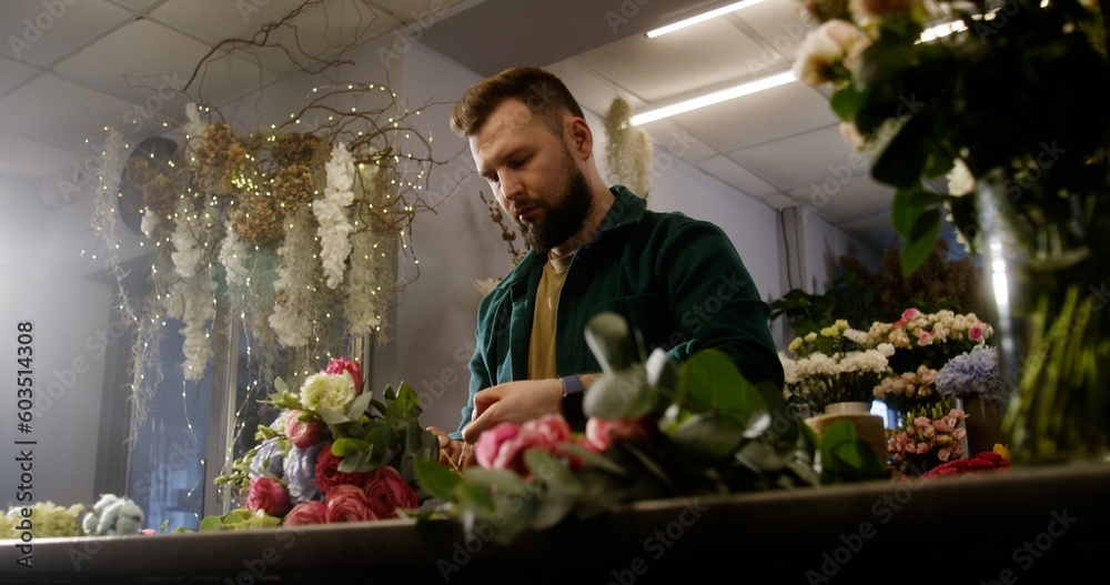 Professional florist entrepreneur ties bunch of fresh flowers using twine. Man collects beautiful bouquet for sale or delivery in flower shop. Concept of retail floral business and entrepreneurship.