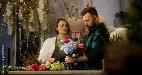 Two professional florists discuss work process and create bouquet in floral shop. Man adds beautiful flowers to bunch. Vases with flowers at background. Concept of floral business and coworking.