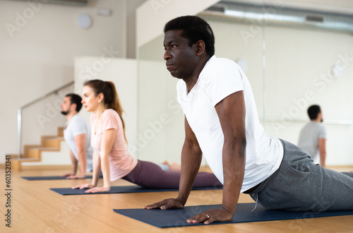 African-american man exercising cobra pose during group yoga training in gym.