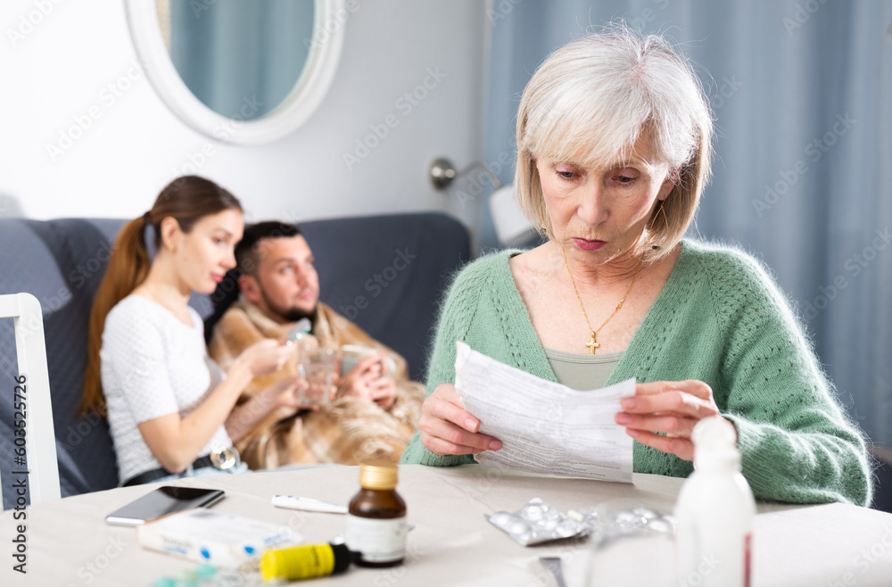Portrait of focused elderly woman sitting at table reading instruction choosing medicines for her sick adult son, while her daughter-in-law trying to help and giving drugs to him