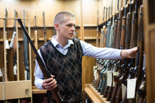 Confident male hunter examining hunting shotgun before buying in gun store..
