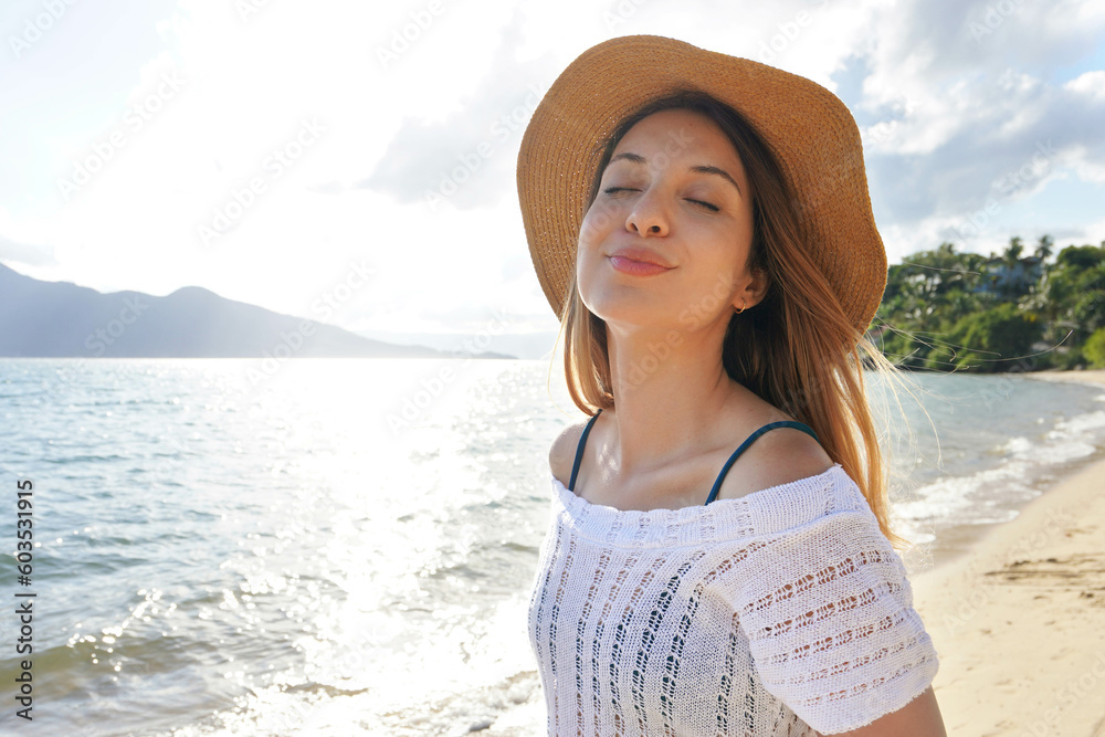 Happy woman with hat breathing and smiling at the beach on summer vacation. Beautiful young female with closed eyes enjoy nature relaxed outdoors. Happy lifestyle and well being concept.