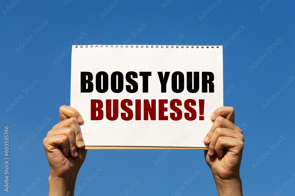 Boost your business text on notebook paper held by 2 hands with isolated blue sky background. This message can be used as business concept about boost your business.