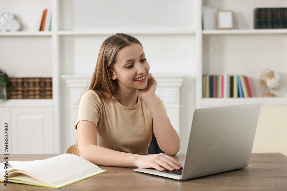 Online learning. Smiling teenage girl with laptop at wooden table indoors Online learning. Smiling teenage girl with laptop at wooden table indoors