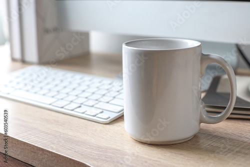 Blank mockup white coffee mug on office table near computer and window with daylight. 