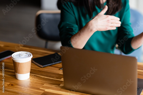 torso of caucasian woman in green dress gesticulating behind laptop with phone and paper to-go cup of coffee on conference table