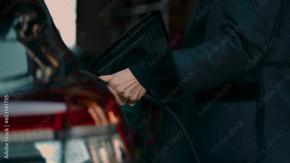 Electric car charging. Young modern woman is connecting plug socket of the home charging station with hand. Black luxurious automobile in a private garage. 4K UHD close up of plugging in power cord.