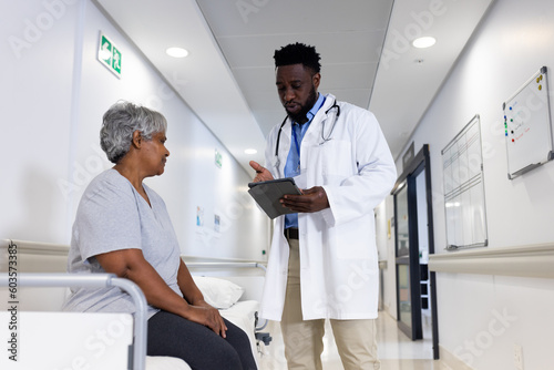 African american male doctor using tablet talking with diverse senior female patient in corridor