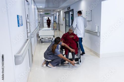 Asian female doctor checking leg of diverse senior female patient in wheelchair in hospital corridor