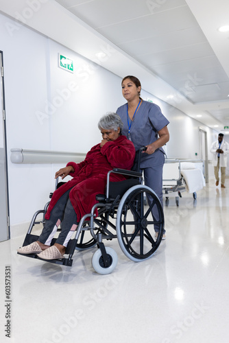 Asian female doctor pushing diverse senior female patient in wheelchair in hospital corridor