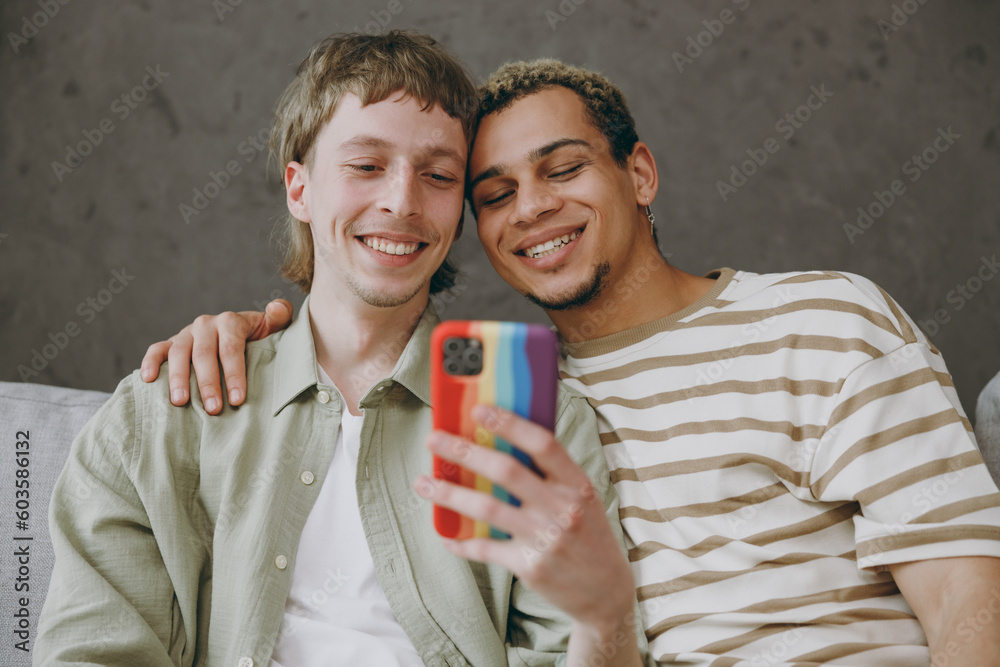 Young happy couple two gay men wear casual clothes together hold use mobile cell phone sit on sofa couch at home flat rest spend free spare time in living room Pride day june month love lgbtq concept - obrazy, fototapety, plakaty 