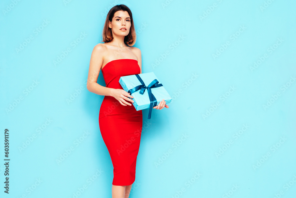 Young beautiful smiling female in red evening dress. Sexy brunette woman isolated on blue. Positive model opens gift box, wondering whats inside as celebrating birthday. Receive b-day presents. Studio Young beautiful smiling female in red evening dress. Sexy brunette woman isolated on blue. Positive model opens gift box, wondering whats inside as celebrating birthday. Receive b-day presents. Studio