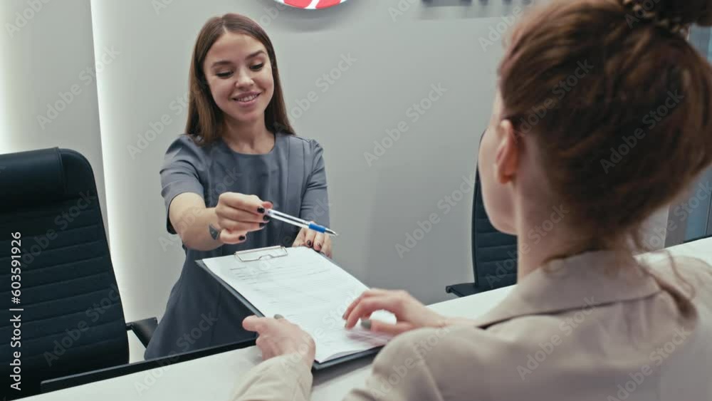 Selective focus shot of young receptionist working at check-in desk in modern clinic explaining patient how to sign document