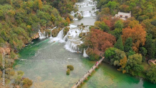 A drone captures the Krka waterfalls in stunning detail. People cross a wooden bridge in the foreground, adding scale and perspective. photo