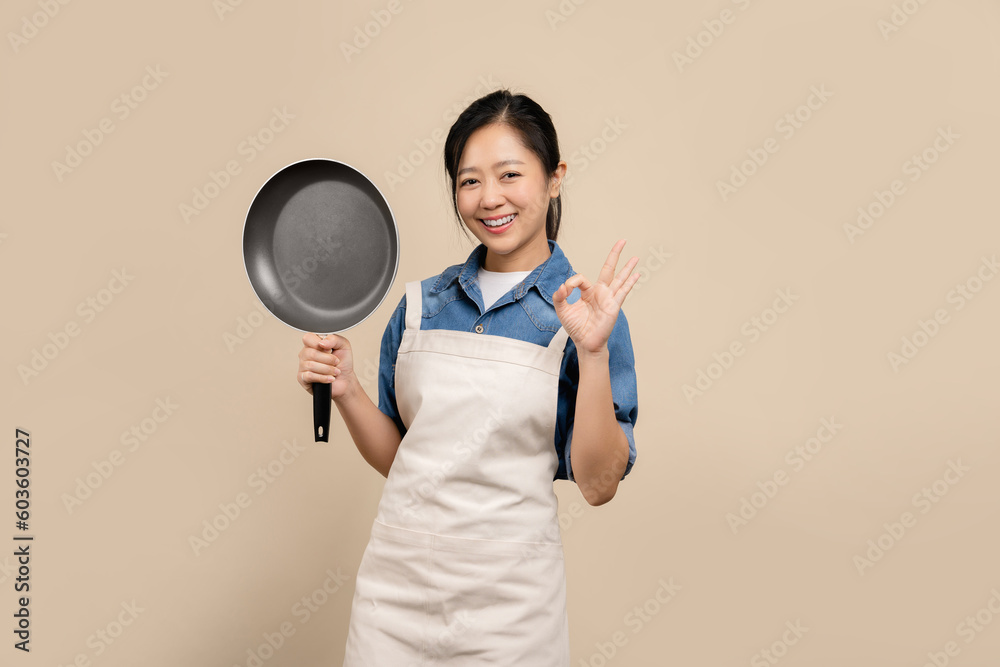 Beautiful Asian woman wearing apron and holding a pan isolated on light brown background. Beautiful Asian woman wearing apron and holding a pan isolated on light brown background.