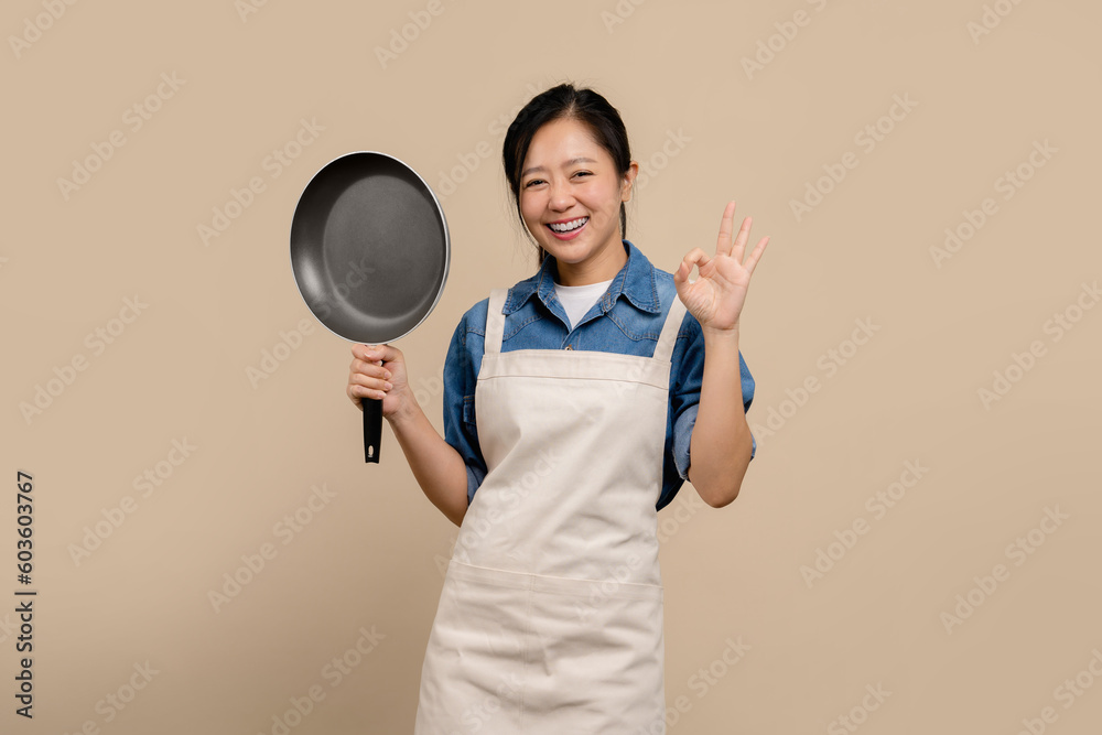 Pretty Asian smiling woman in apron and holding frying pan standing isolated on light brown background. She is doing OK gesture. Pretty Asian smiling woman in apron and holding frying pan standing isolated on light brown background. She is doing OK gesture.