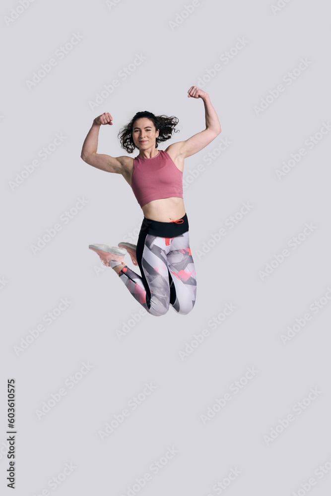 Determined strong sportswoman showing biceps muscles, jumping high up, isolated on white background