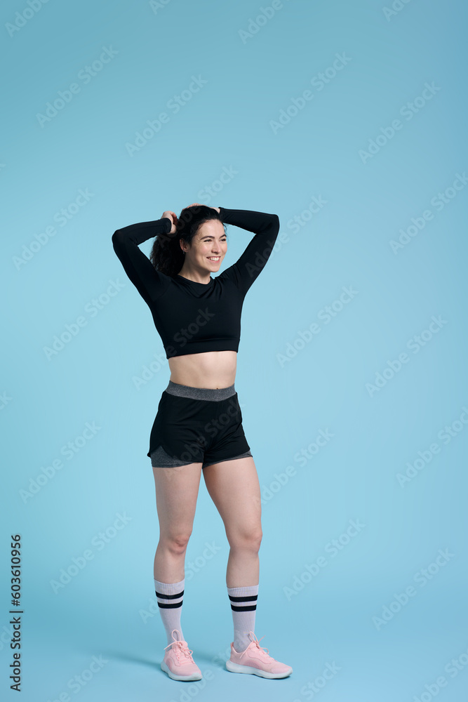Young smiling strong fitness woman wearing black bra and sports shorts, standing over blue backdrop Young smiling strong fitness woman wearing black bra and sports shorts, standing over blue backdrop