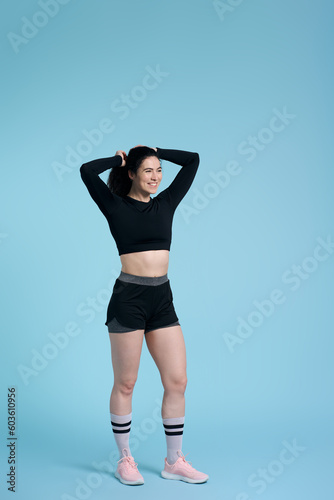 Young smiling strong fitness woman wearing black bra and sports shorts, standing over blue backdrop