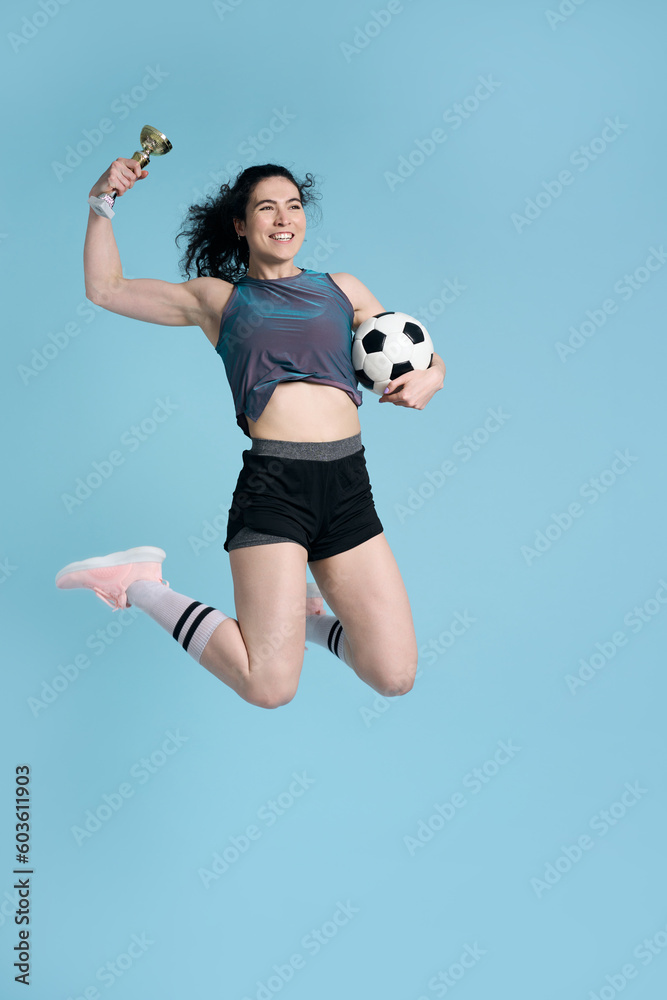 Fototapeta premium Athlete woman smiles, jumps high up over blue background, holds soccer ball and golden champion cup